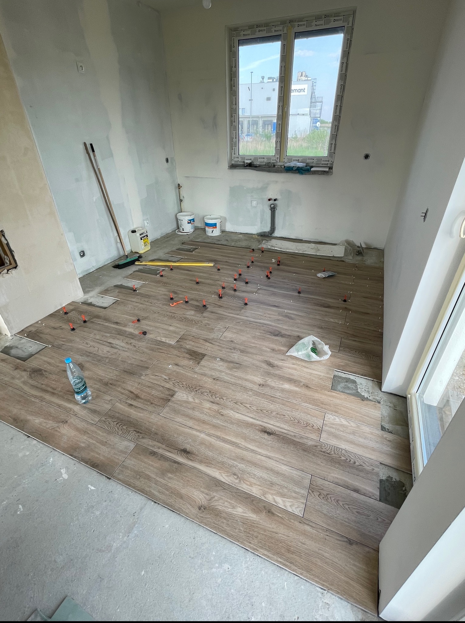 Układanie podłogi z płytek imitujących drewno w jasnym wnętrzu. Widoczne poziomica, butelka wody i system poziomowania płytek. Częściowo wykończone ściany.