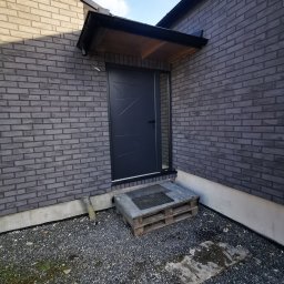 Adrian Kurczewski - Eleganckie wejście do domu z szarej cegły, zadaszone drewnianym daszkiem i nowoczesnymi, czarnymi drzwiami z geometrycznym wzorem. Żwirowe podłoże.