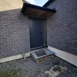 Eleganckie wejście do domu z szarej cegły, zadaszone drewnianym daszkiem i nowoczesnymi, czarnymi drzwiami z geometrycznym wzorem. Żwirowe podłoże.