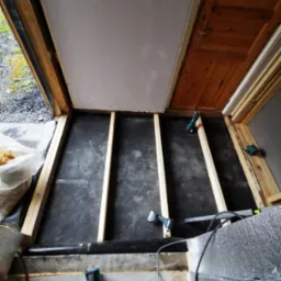 Montaż drewnianej podłogi na czarnej membranie, widoczne legary i narzędzia. Etap pośredni prac wykończeniowych, z perspektywy podłogi, z fragmentem drzwi i ścian.