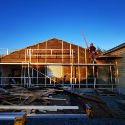 Adrian Kurczewski - Montaż izolacji budynku szkieletowego z rusztowania. Widoczna piła DeWalt i deski. Niebieskie niebo w tle. Praca w toku.