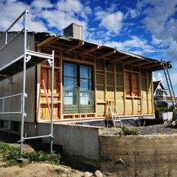 Adrian Kurczewski - Budowa domu szkieletowego w trakcie: widoczne rusztowania, drewniana konstrukcja ścian z izolacją, okno i drabina. Ujęcie z dołu, błękitne niebo w tle.