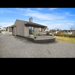 Adrian Kurczewski - Nowoczesny dom z szarej cegły z płaskim dachem i kominem. Widoczny taras z meblami ogrodowymi i szara nawierzchnia. Błękitne niebo w tle.