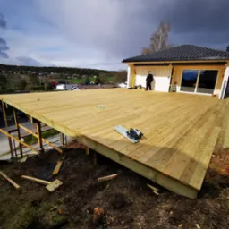 Montaż tarasu drewnianego przy domu jednorodzinnym. Widoczne narzędzia pracy, częściowo ukończona konstrukcja i osoba w tle. Pochmurne niebo.