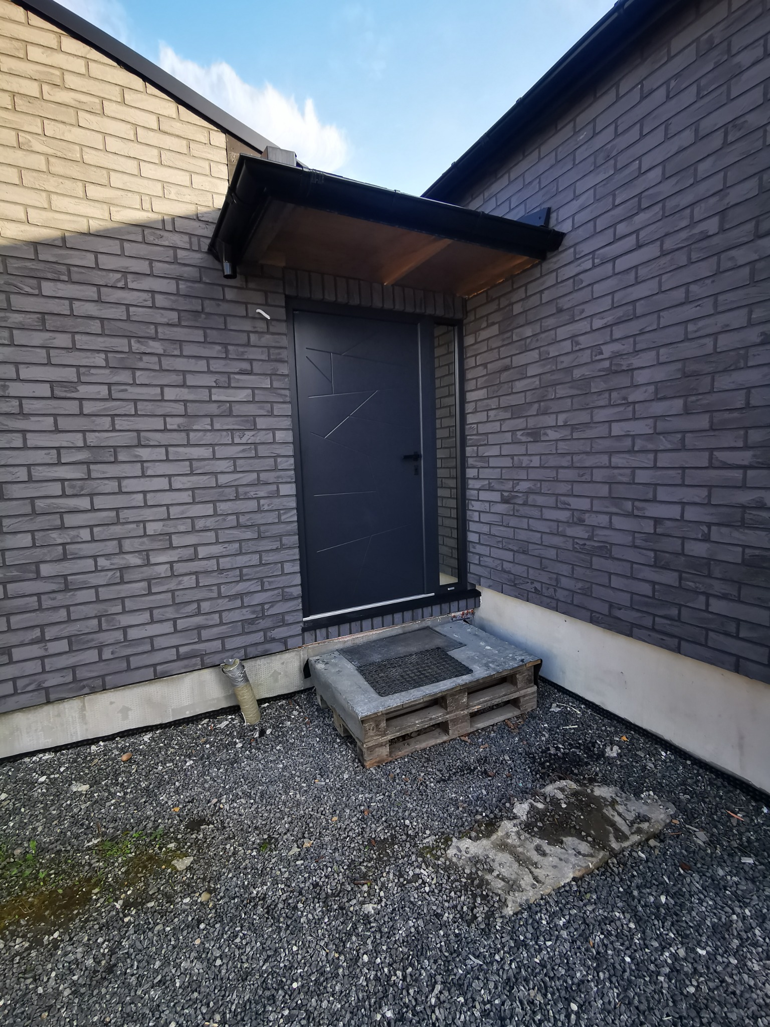 Eleganckie wejście do domu z szarej cegły, zadaszone drewnianym daszkiem i nowoczesnymi, czarnymi drzwiami z geometrycznym wzorem. Żwirowe podłoże.