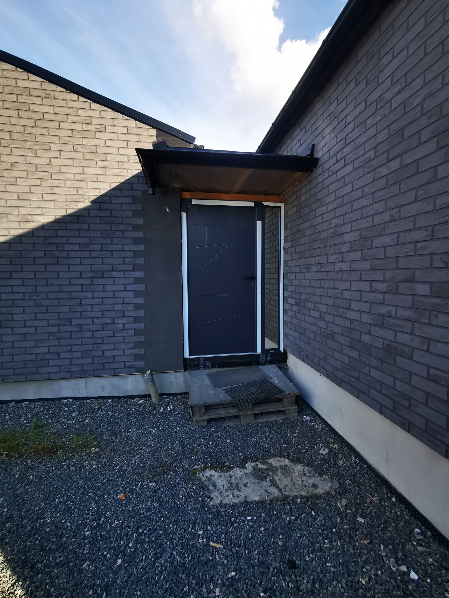 Nowoczesne wejście do domu z ciemnej cegły, z czarnymi drzwiami o geometrycznym wzorze i zadaszeniem. Podłoże wyłożone drobnym, ciemnym żwirem. Minimalistyczny design.