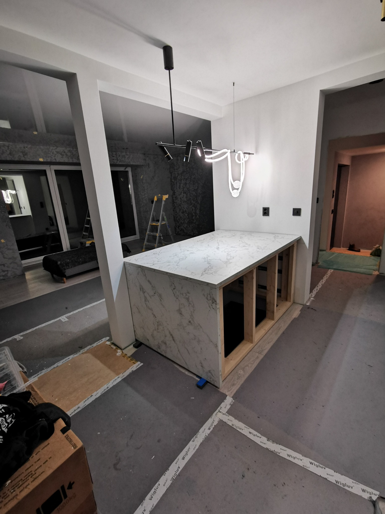 Wnętrze w trakcie wykańczania: wyspa kuchenna z blatem imitującym marmur na drewnianej konstrukcji, nowoczesne oświetlenie, widoczne materiały budowlane.