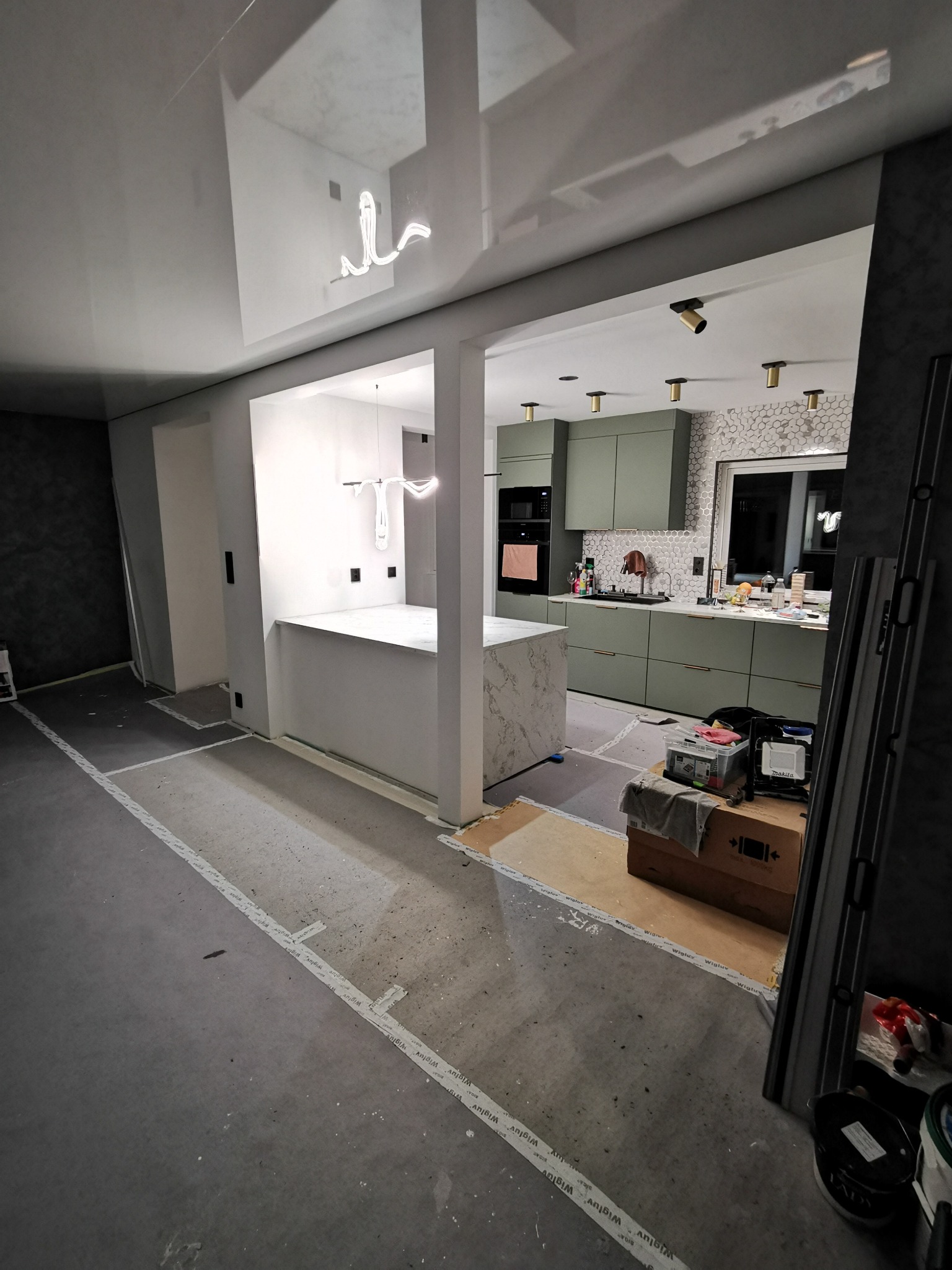 Wnętrze w trakcie wykończenia: surowa podłoga z taśmami, widoczna kuchnia z zielonymi szafkami i marmurowym blatem, sufit z nowoczesnym oświetleniem. Prace wykończeniowe w toku.