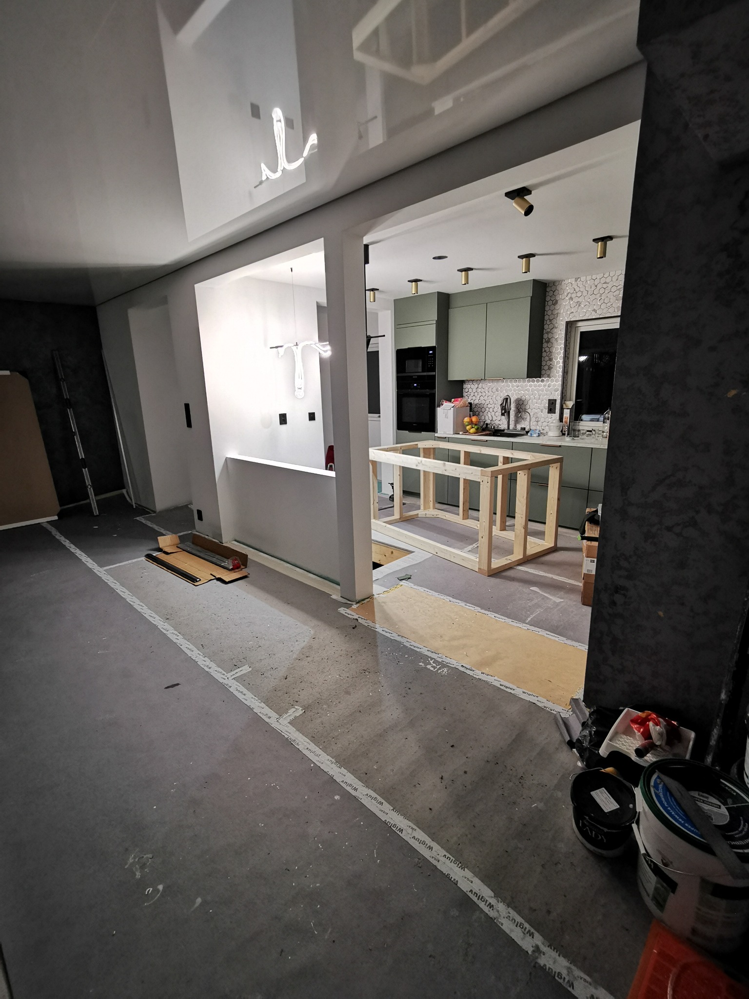 Wnętrze w trakcie wykończenia w Dalikowie: widok na kuchnię z zabudową w odcieniach zieleni, drewniana konstrukcja wyspy, surowa podłoga i elementy oświetlenia.