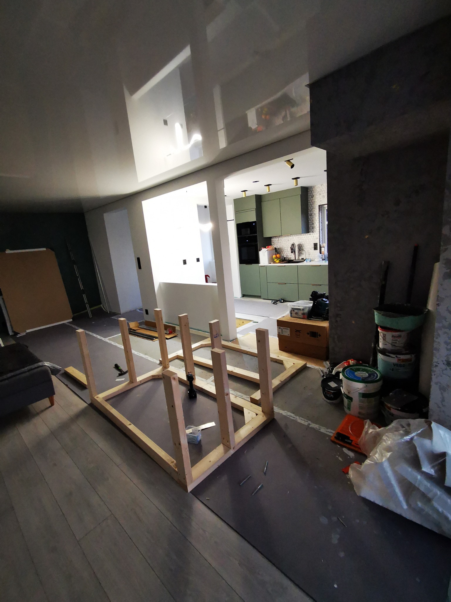 Wnętrze w trakcie remontu: drewniana konstrukcja podłogi na tle szarej podłogi, widoczna kuchnia w głębi. Puszki z farbą i narzędzia obok. Odblaski na suficie.