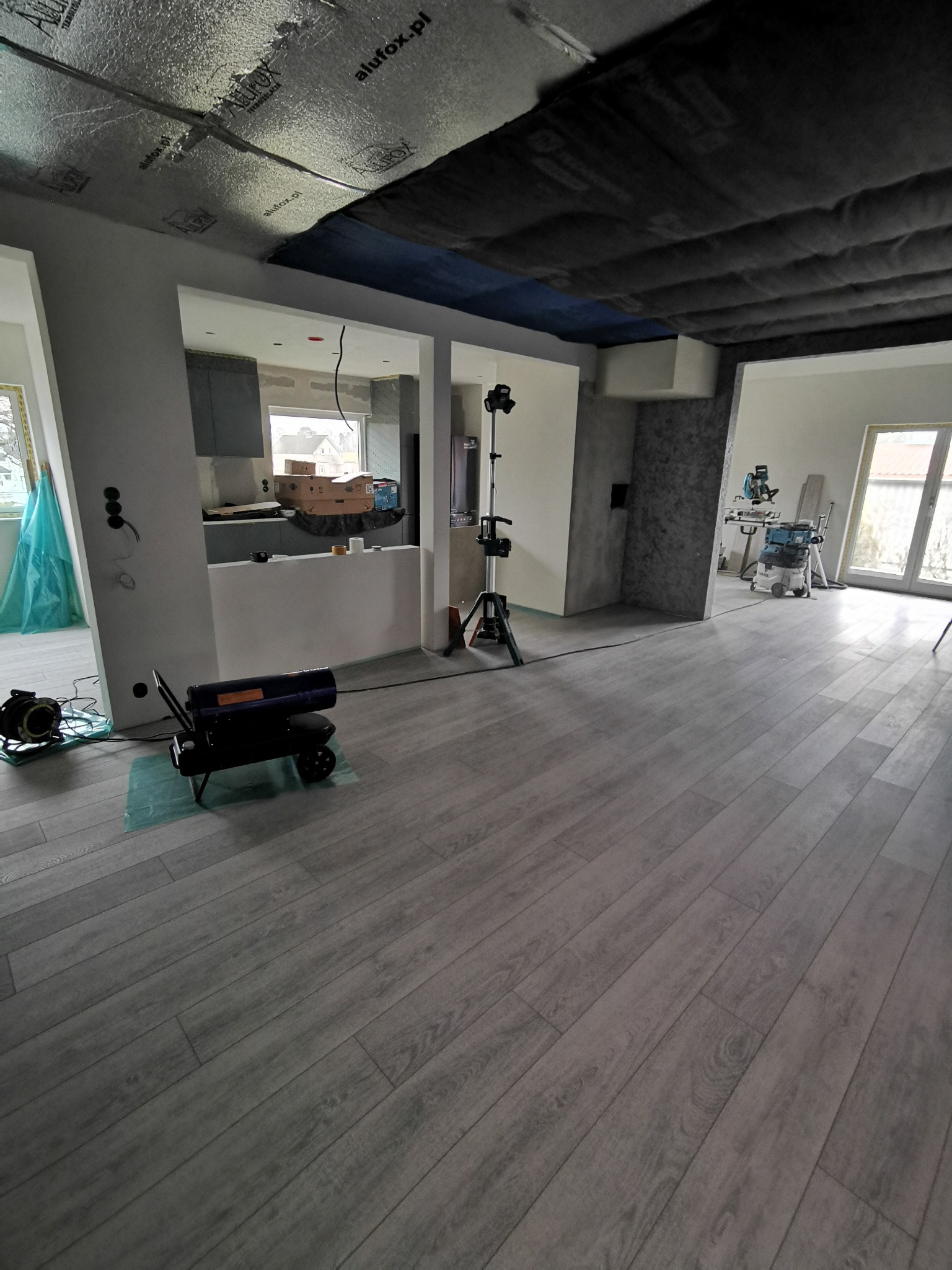 Wnętrze w trakcie wykończenia: szara podłoga, widoczne narzędzia (nagrzewnica, lampa), sufit z izolacją. Częściowo wykończona kuchnia w tle.