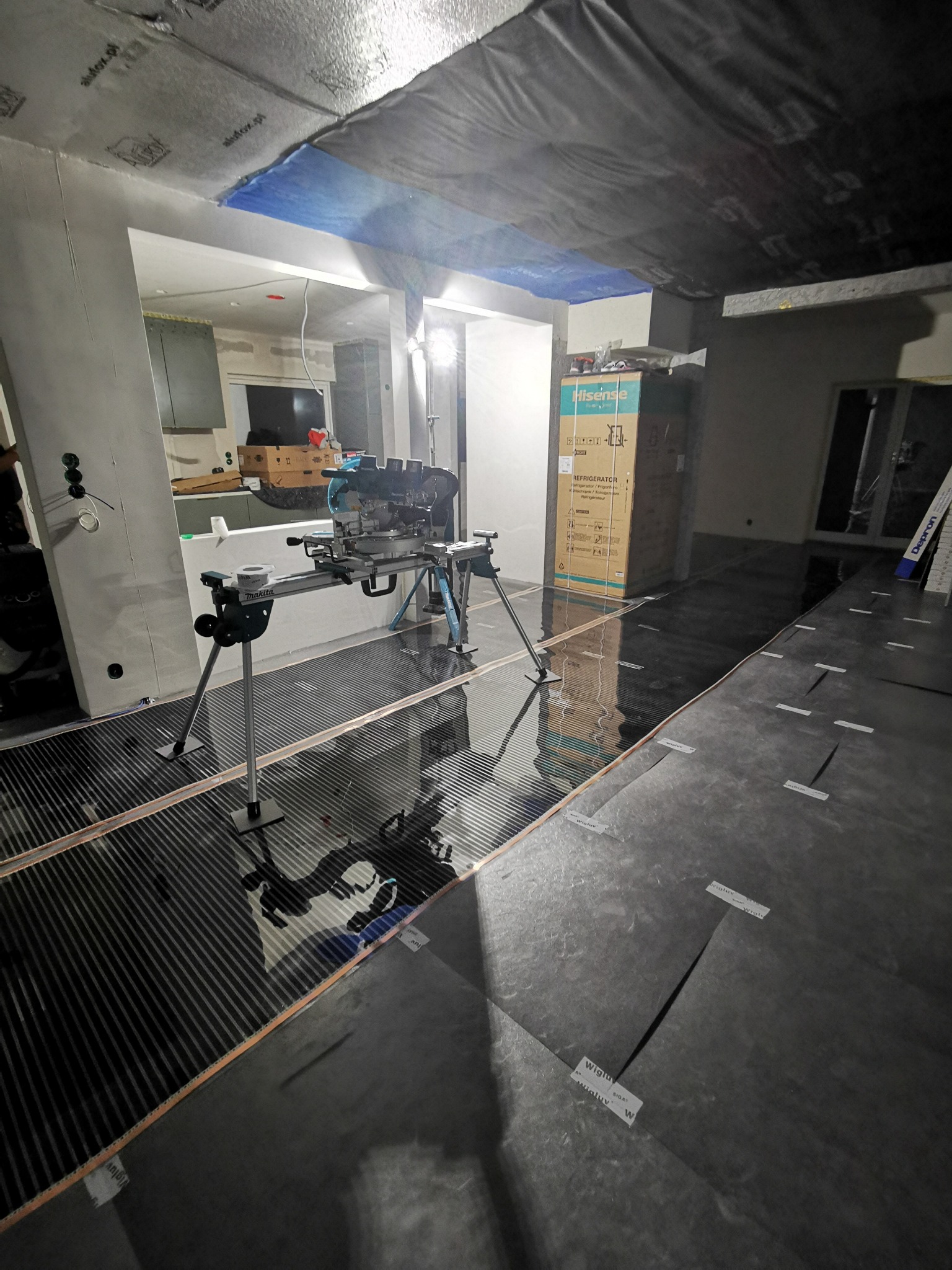 Instalacja folii grzewczej na podłodze w trakcie remontu. Widoczny sprzęt do cięcia folii. W tle karton z lodówką. Oświetlenie górne w folii aluminiowej.