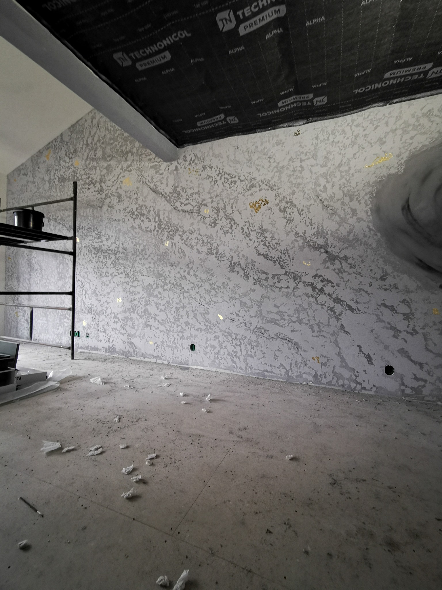 Wykończona ściana z dekoracyjnym tynkiem w odcieniach szarości z złotymi akcentami, widoczna w surowym wnętrzu w trakcie prac wykończeniowych.