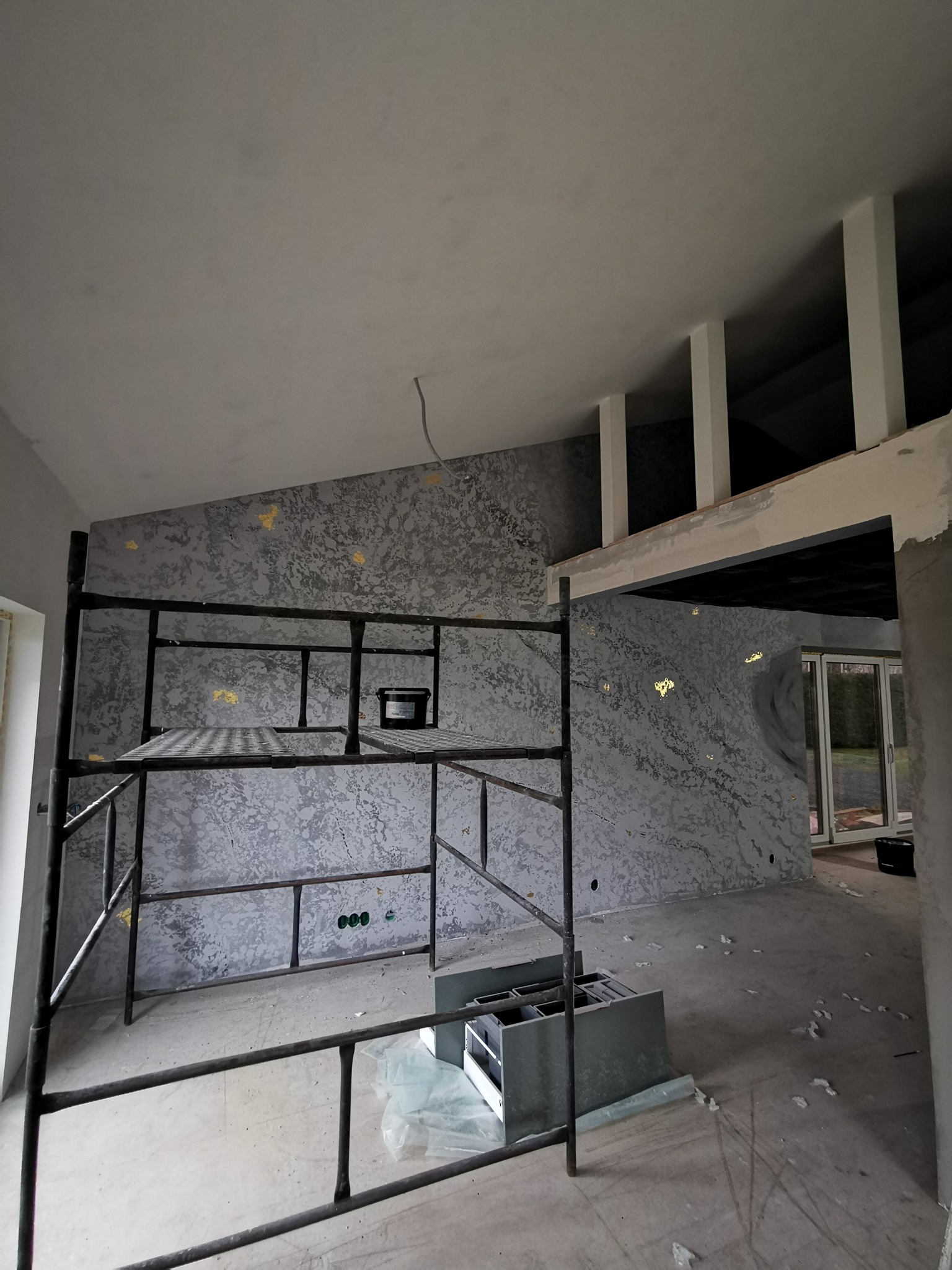 Ściana z dekoracyjnym tynkiem w odcieniach szarości i złotymi akcentami, widoczna z rusztowania w trakcie prac wykończeniowych wnętrza. Widoczne puszki elektryczne.