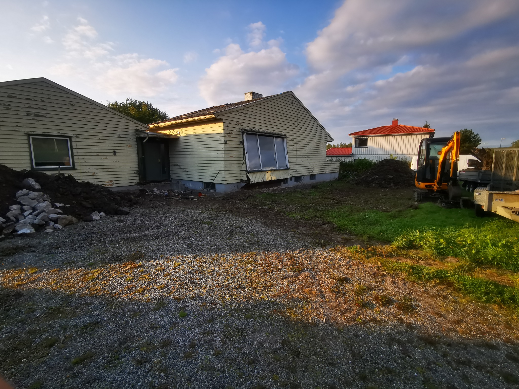 Dom z żółtą elewacją w trakcie prac ziemnych. Na pierwszym planie żwir, w tle koparka i przyczepa. Widoczne ślady demontażu elementów elewacji.