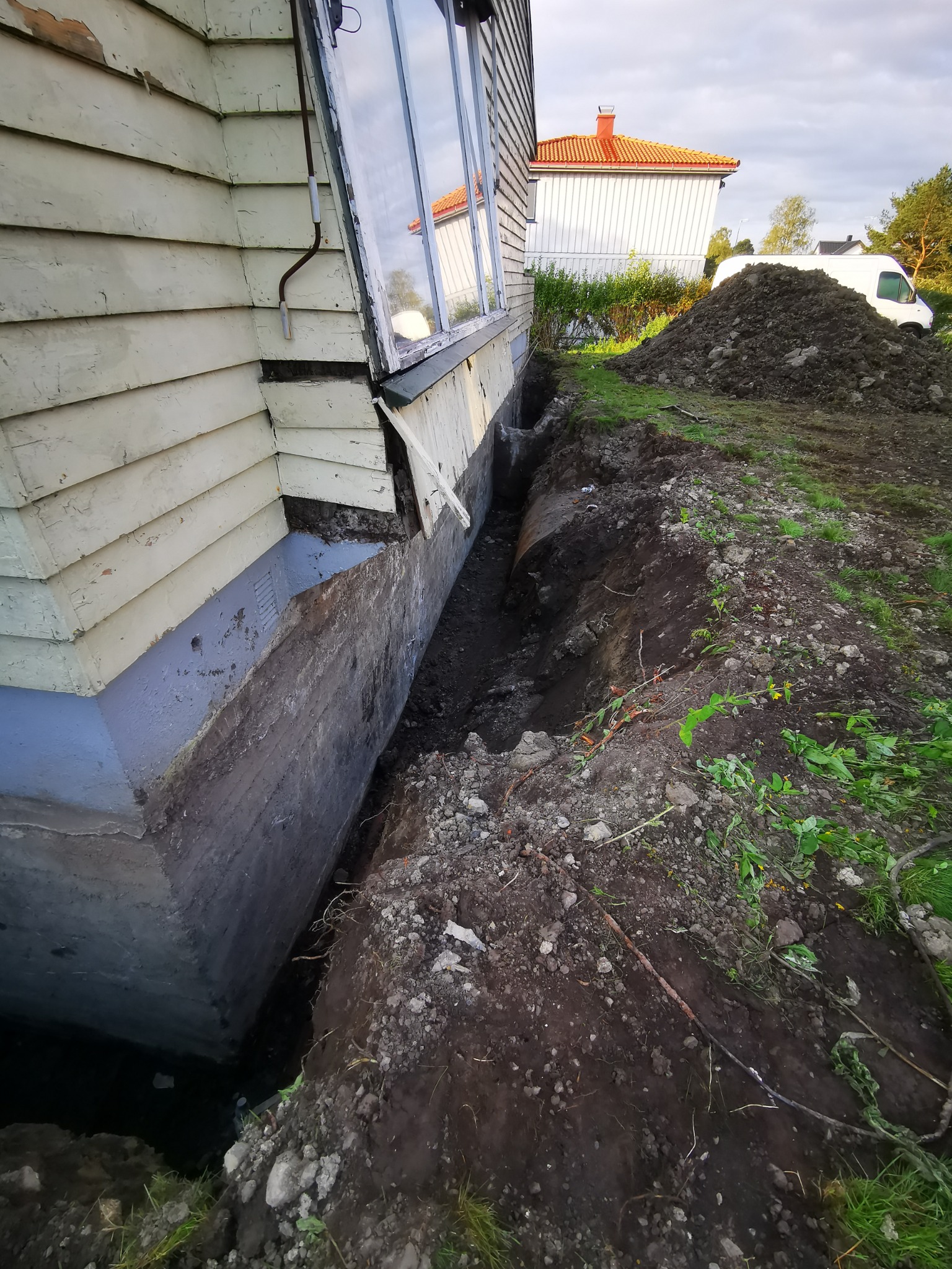 Odkopany fundament drewnianego domu z oknem, przygotowany do prac izolacyjnych. W tle hałda ziemi i biały samochód dostawczy.