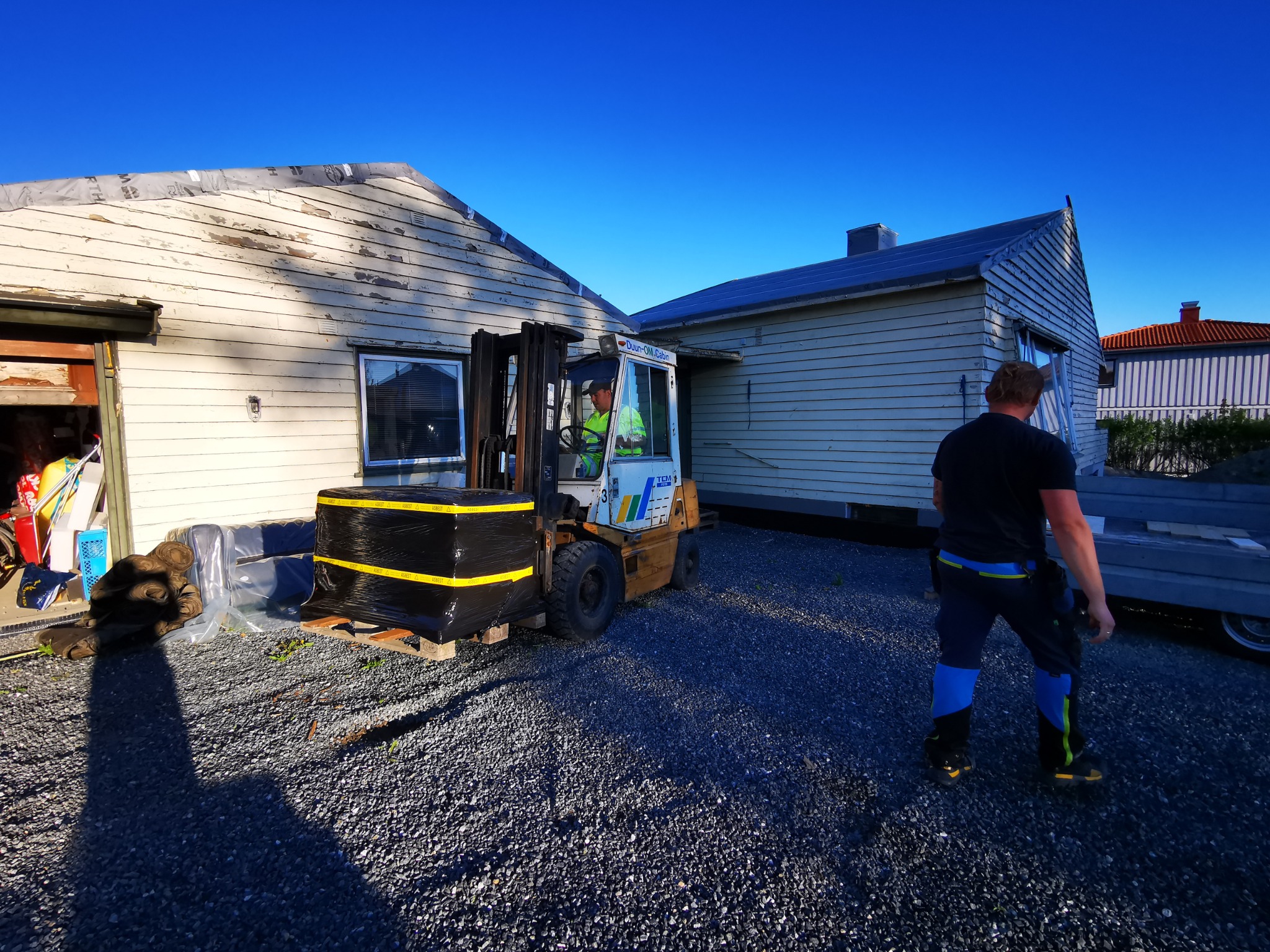 Montaż dachu: wózek widłowy z materiałami budowlanymi na tle domu z częściowo zdjętym poszyciem. Pracownik w oddali.