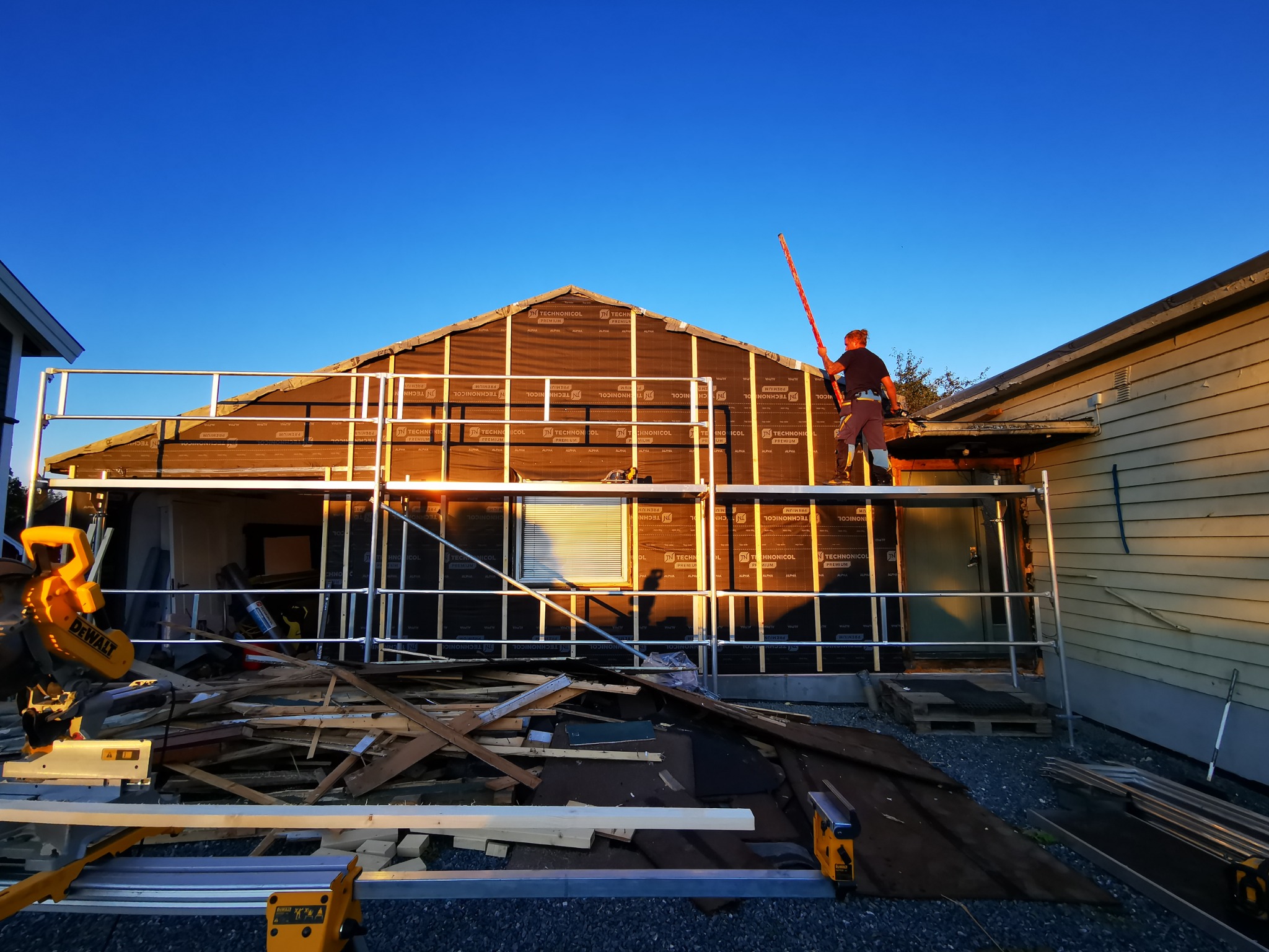 Montaż izolacji budynku szkieletowego z rusztowania. Widoczna piła DeWalt i deski. Niebieskie niebo w tle. Praca w toku.