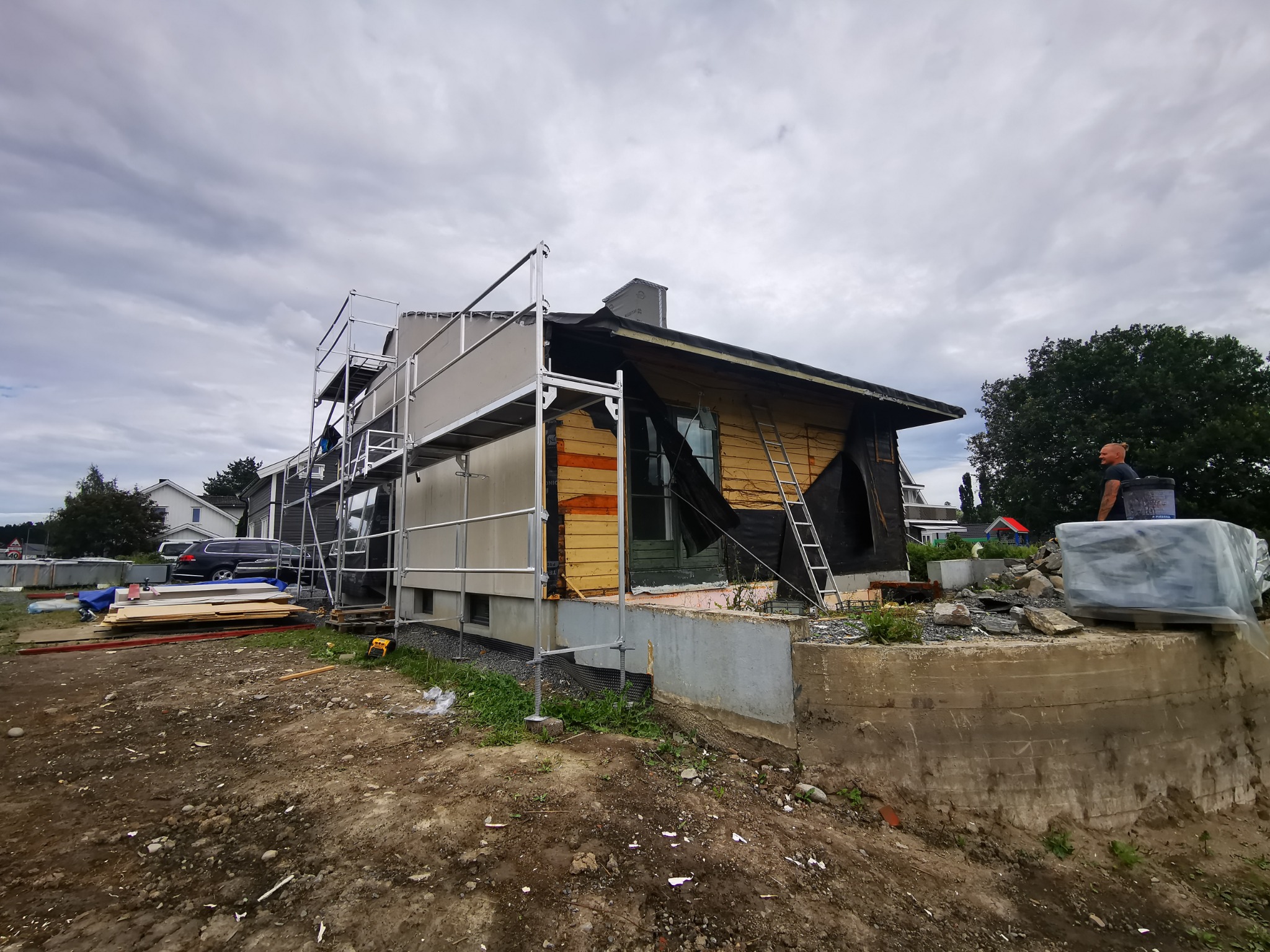 Dom w trakcie remontu elewacji, widoczne rusztowania, odkryte deski i częściowo położona izolacja. Na pierwszym planie materiały budowlane i teren wokół domu.