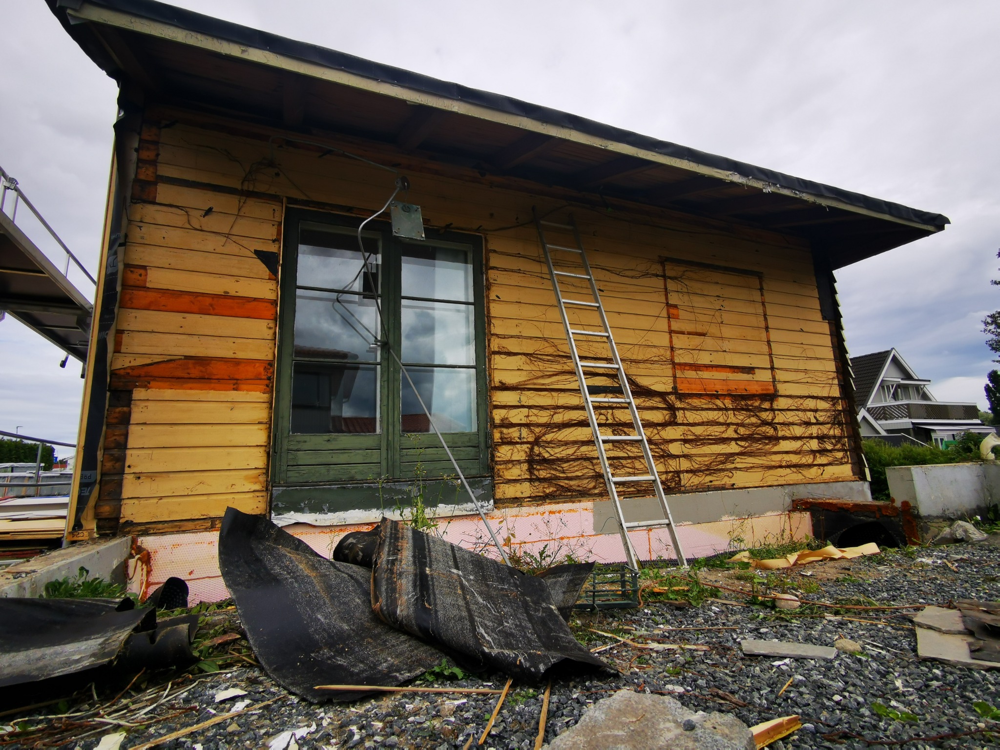 Budynek w trakcie remontu elewacji z widoczną drabiną, odsłoniętą izolacją i resztkami materiałów budowlanych na ziemi. Widoczne drewniane deski i okno.