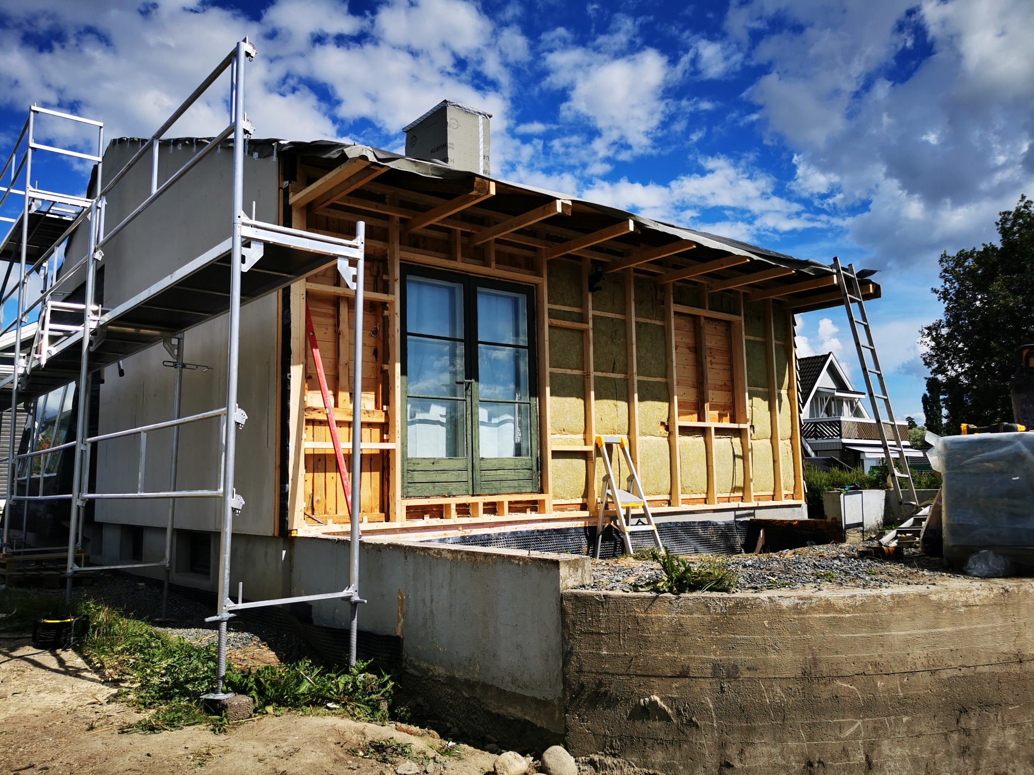 Budowa domu szkieletowego w trakcie: widoczne rusztowania, drewniana konstrukcja ścian z izolacją, okno i drabina. Ujęcie z dołu, błękitne niebo w tle.