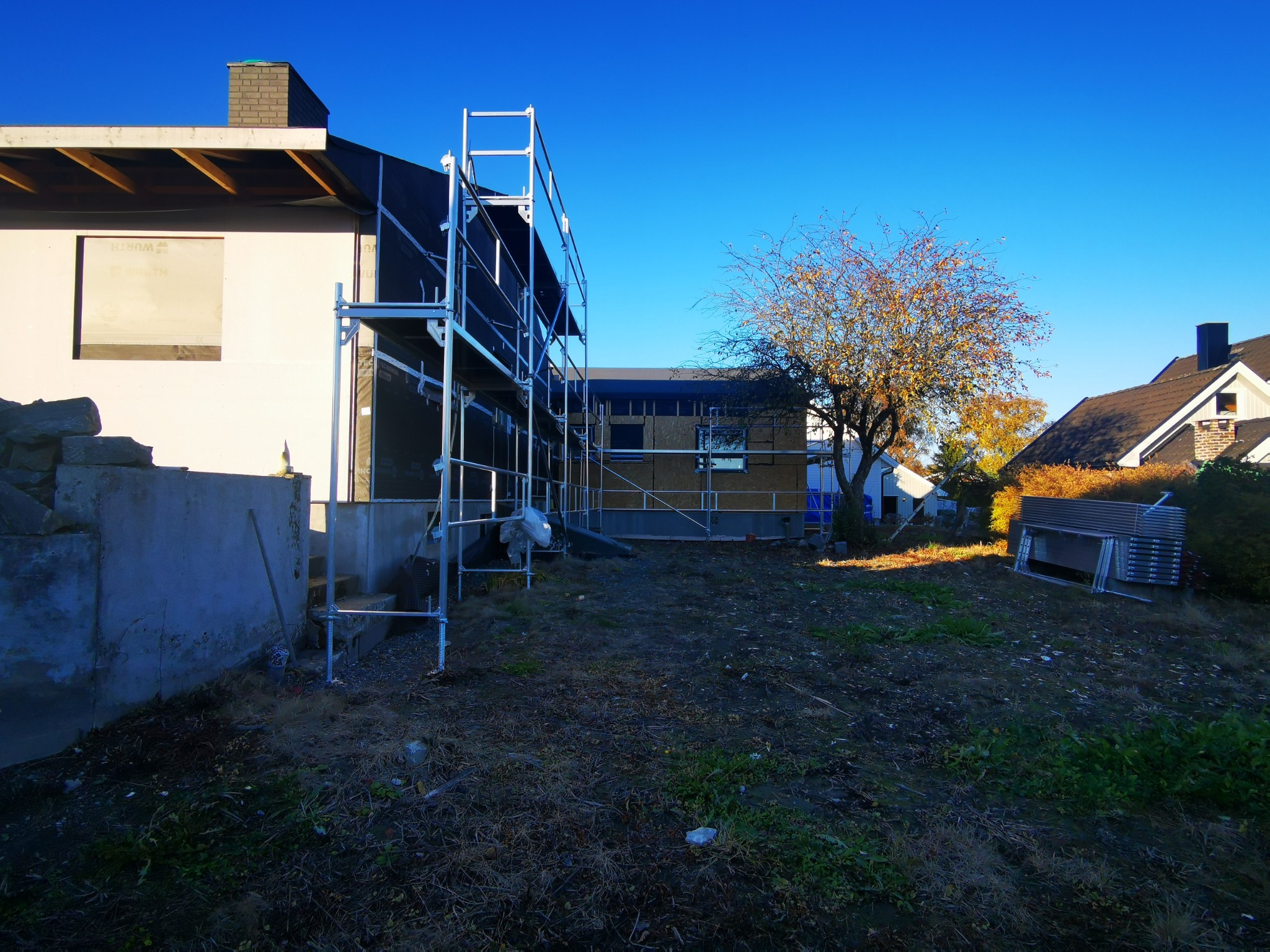 Budowa domu szkieletowego w toku: widoczne rusztowania, fragmenty konstrukcji i ocieplenia na tle błękitnego nieba. Prace ziemne w otoczeniu budynku.