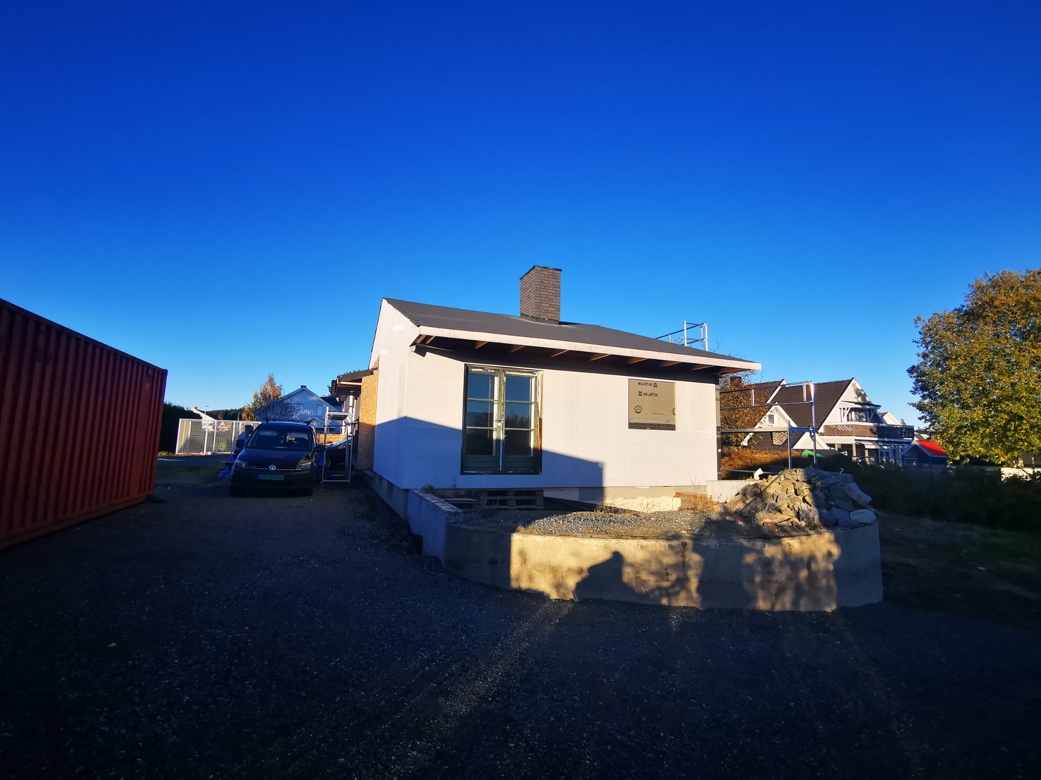 Niedokończony dom szkieletowy z widocznym kominem i oknem, obok zaparkowany samochód i kontener, w tle inne domy i błękitne niebo.