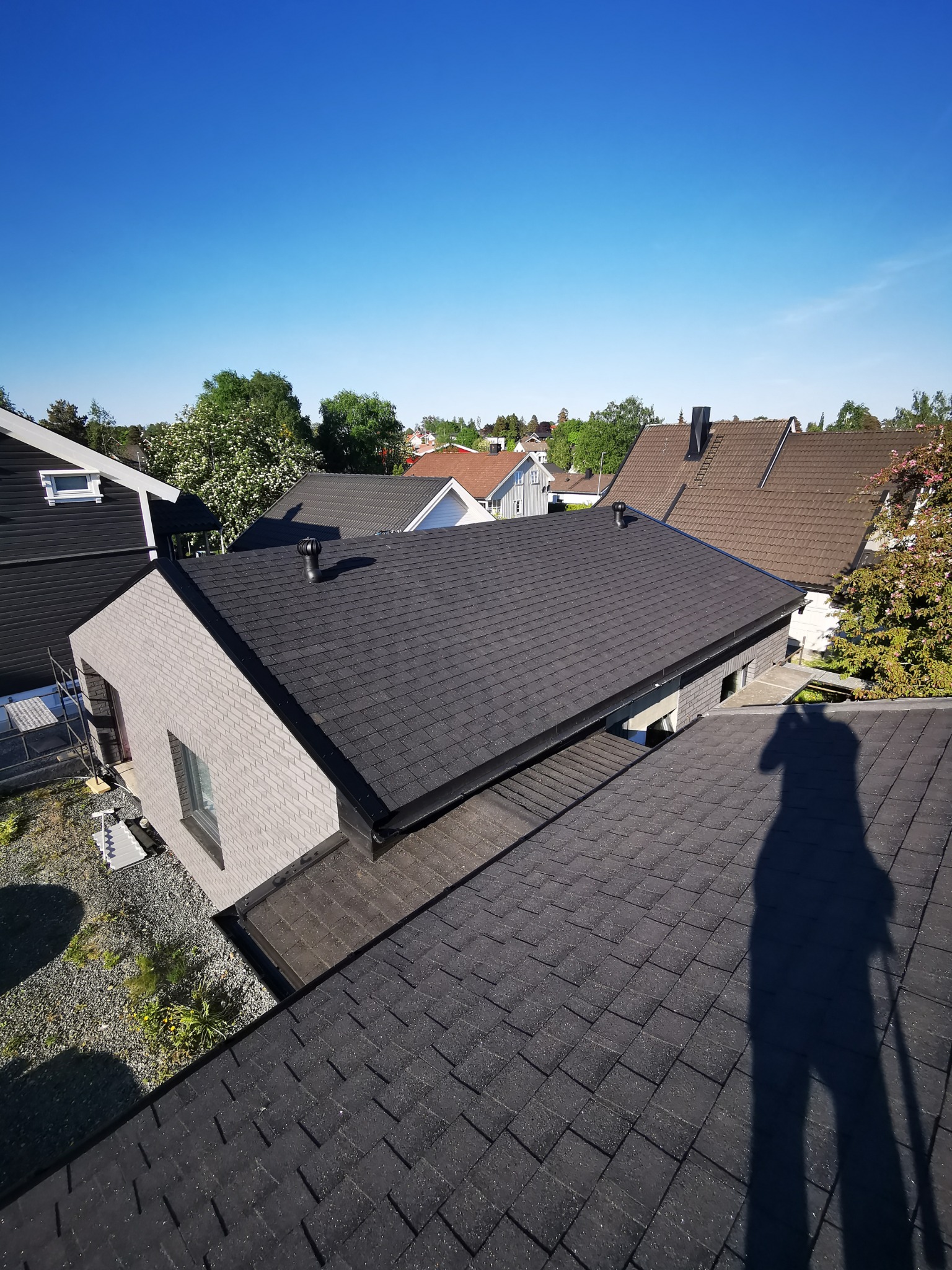 Widok z góry na nowo pokryty dach domu z bitumicznych gontów, z widocznym cieniem osoby robiącej zdjęcie, otoczonego innymi budynkami i zielenią w słoneczny dzień.