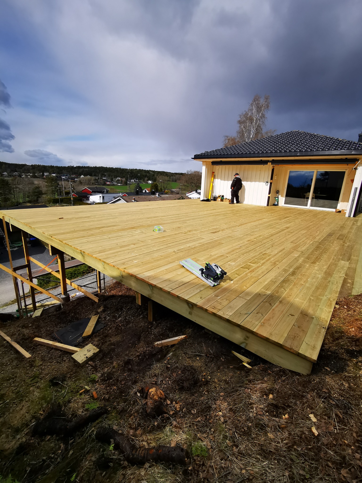 Montaż tarasu drewnianego przy domu jednorodzinnym. Widoczne narzędzia pracy, częściowo ukończona konstrukcja i osoba w tle. Pochmurne niebo.