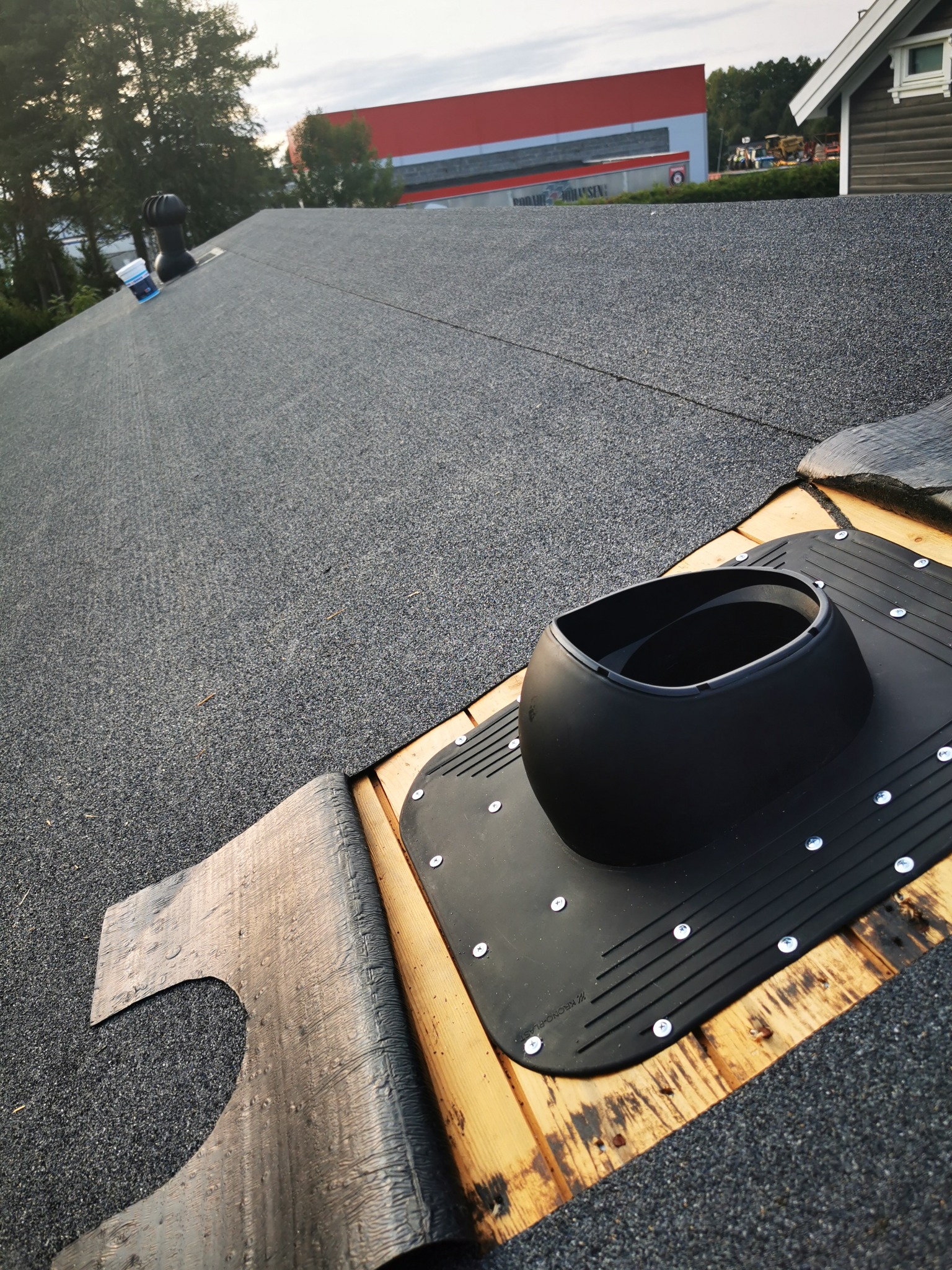 Szczegółowe ujęcie fragmentu dachu z papy z zamontowanym kominkiem wentylacyjnym. Widoczne drewniane elementy konstrukcyjne i faktura pokrycia dachowego.