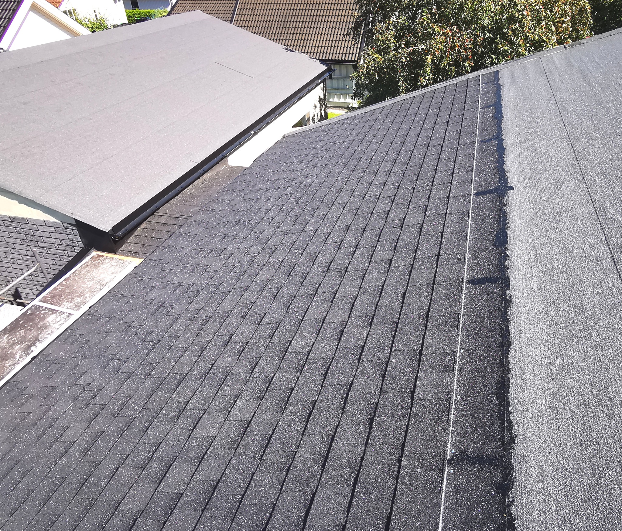 Fragment dachu z gontów bitumicznych i papy termozgrzewalnej. Widoczne połączenie dwóch różnych typów pokrycia dachowego. Ujęcie z góry, ostre światło słoneczne.