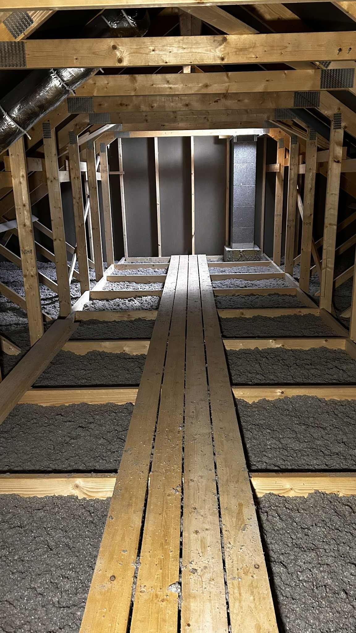 Poddasze z drewnianą konstrukcją i szarą izolacją w przestrzeniach między belkami, widoczna kładka. Wentylacja w rogu.