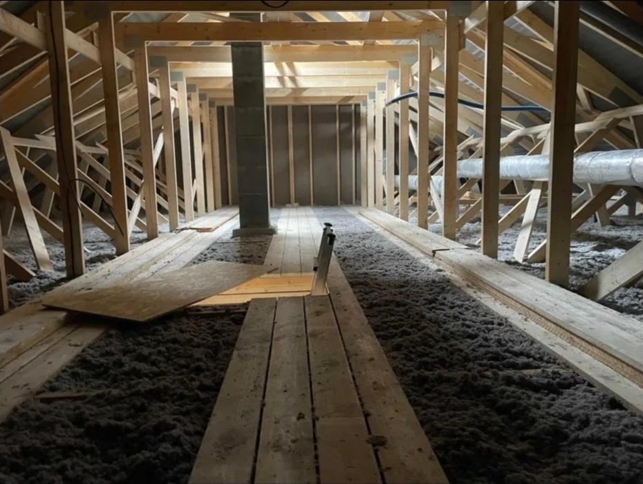 Poddasze z widoczną konstrukcją drewnianą i warstwą izolacji z wełny mineralnej, tymczasowe deski ułożone dla dostępu, szara izolacja wypełnia przestrzeń.