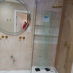 Bud Pro Yevhenii Hlukhenko - Nowoczesna łazienka z okrągłym lustrem z podświetleniem, złotą armaturą i kabiną prysznicową typu walk-in. Widoczne białe marmurowe płytki na podłodze.