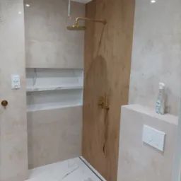 Wykończona kabina prysznicowa z złotą armaturą i półkami w Łodzi. Beżowe płytki, odpływ liniowy, nowoczesny design. Umywalka częściowo widoczna.