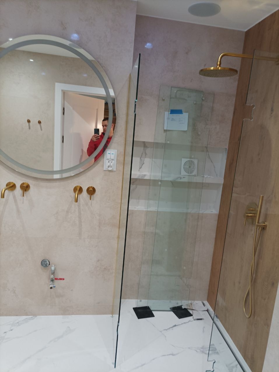 Nowoczesna łazienka z okrągłym lustrem z podświetleniem, złotą armaturą i kabiną prysznicową typu walk-in. Widoczne białe marmurowe płytki na podłodze.