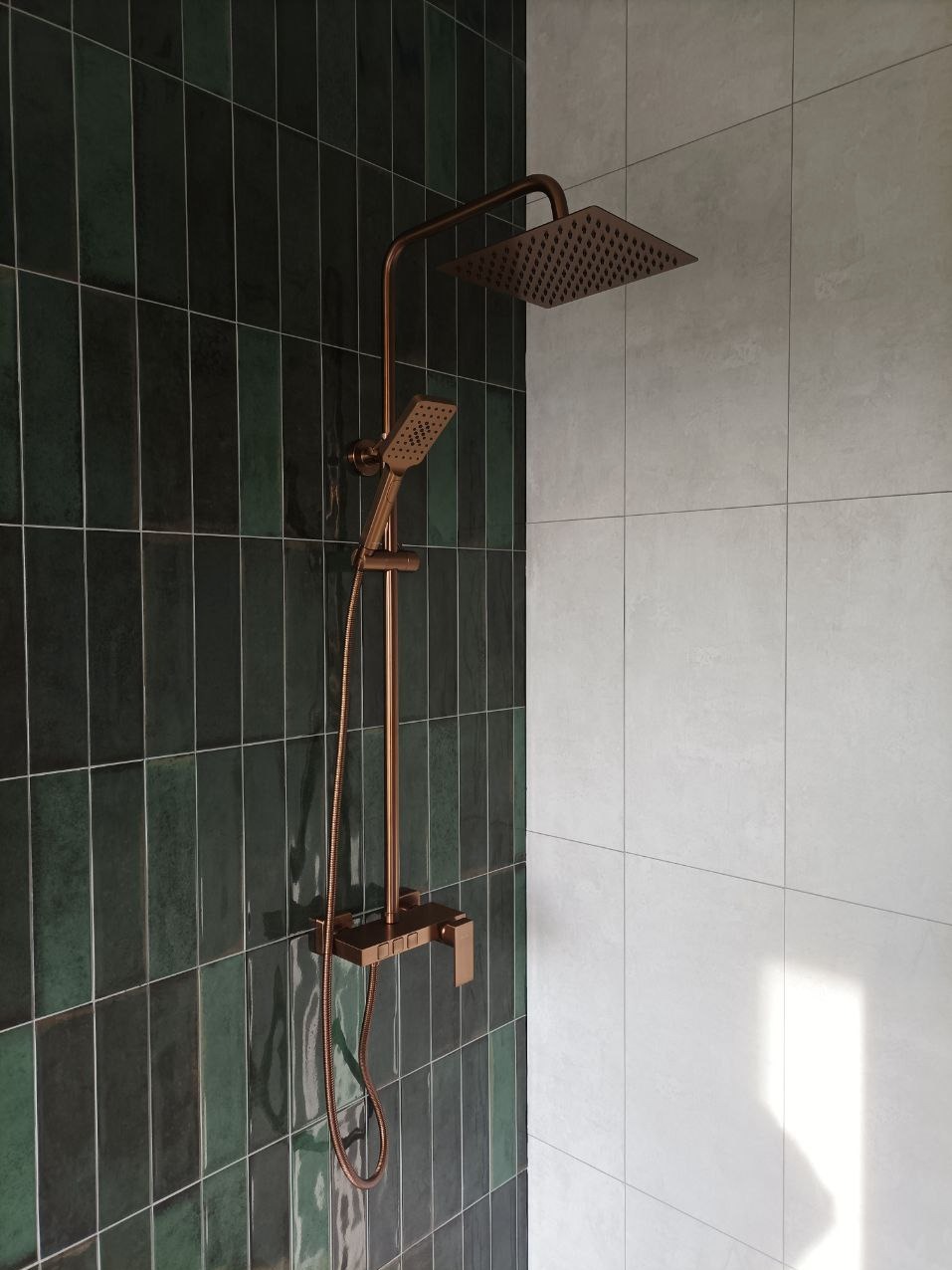 Nowoczesna kabina prysznicowa z baterią w kolorze miedzi, na tle zielonych, pionowych płytek i jasnych, kwadratowych płytek. Minimalistyczny design.