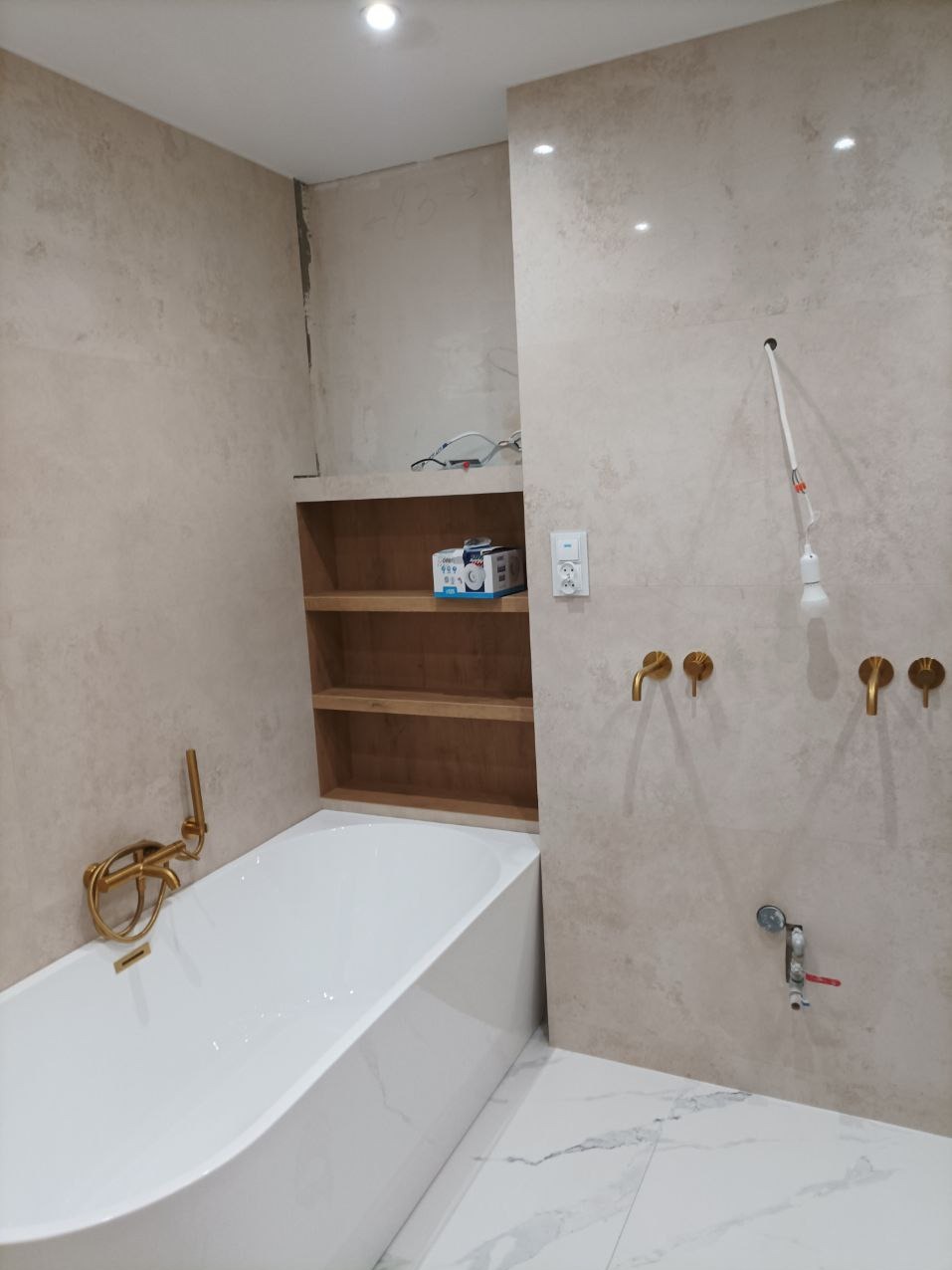 Łazienka w trakcie wykończenia: biała wanna z złotą armaturą, półki w ścianie, płytki w beżowym odcieniu, widoczne elementy instalacji przed ostatecznym montażem.