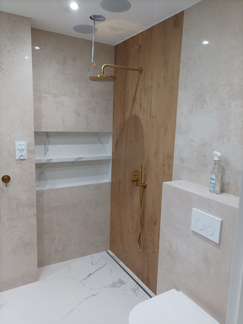 Wykończona kabina prysznicowa z złotą armaturą i półkami w Łodzi. Beżowe płytki, odpływ liniowy, nowoczesny design. Umywalka częściowo widoczna.