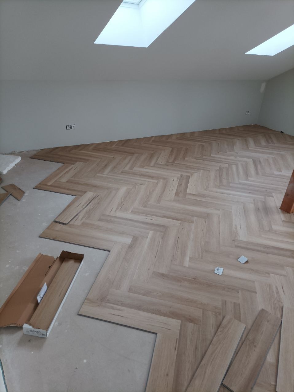 Układanie jasnych paneli podłogowych w jodełkę francuską, widoczne puste kartony i niedokończony fragment podłogi. W tle ściana i okna dachowe.