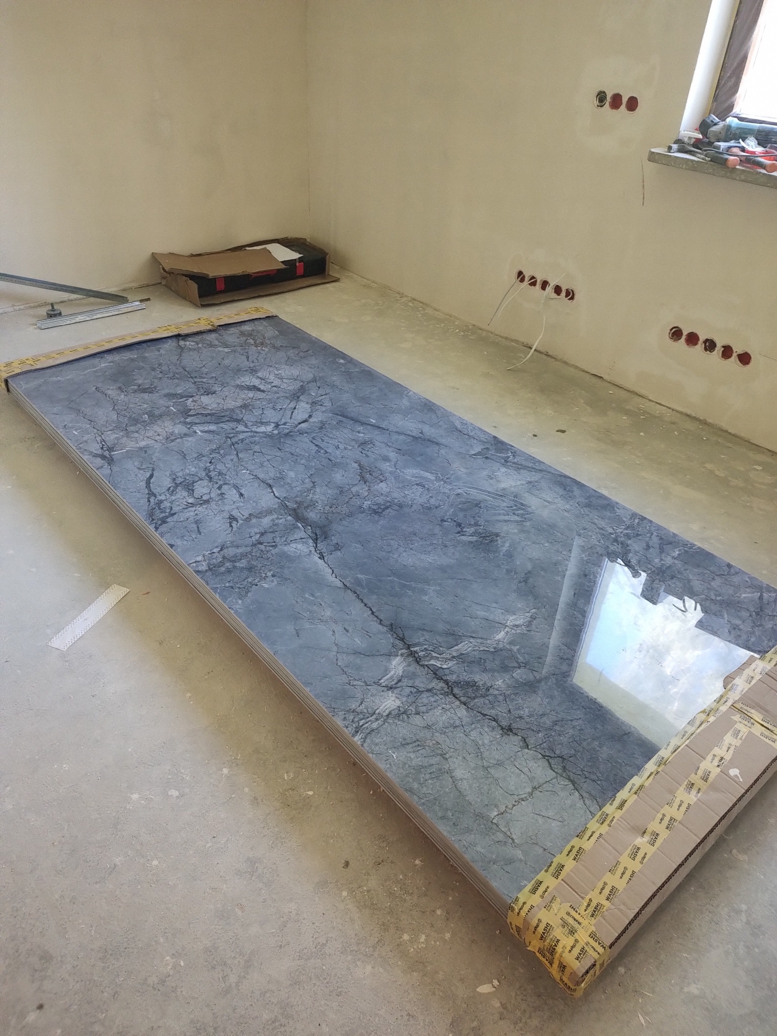 Płyta gresowa w odcieniach szarości z marmurkowym wzorem, zabezpieczona kartonem, leży na podłodze w surowym wnętrzu. Widoczne puszki elektryczne i narzędzia.