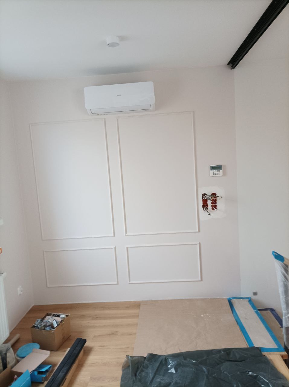 Pokój z dekoracyjną sztukaterią na ścianie, klimatyzatorem i widocznymi przewodami elektrycznymi przed wykończeniem. Na podłodze zabezpieczenia.