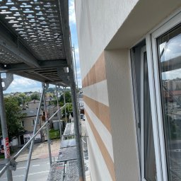 Lichocki patryk - Elewacja budynku w kremowych pasach, widziana ze rusztowania z perspektywy góry. Okno z białymi ramami. W tle zabudowa miejska i zieleń.