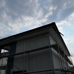 Lichocki patryk - Dom w trakcie tynkowania elewacji, widoczne rusztowania. Częściowo otynkowana ściana budynku na tle pochmurnego nieba. Detal: rynna przy dachu.