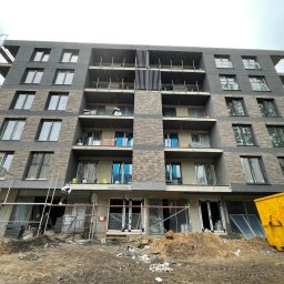 Lichocki patryk - Wielopiętrowy budynek z elewacją w trakcie realizacji. Widoczne rusztowania, pracownicy i kontener na odpady. Część okien zabezpieczona folią.