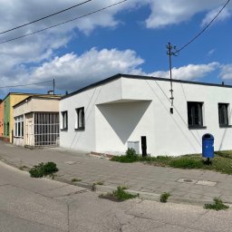 Lichocki patryk - Biały, odnowiony budynek z ciemnymi oknami i niebieskim koszem na śmieci na chodniku, kontrastujący z błękitnym niebem i chmurami.