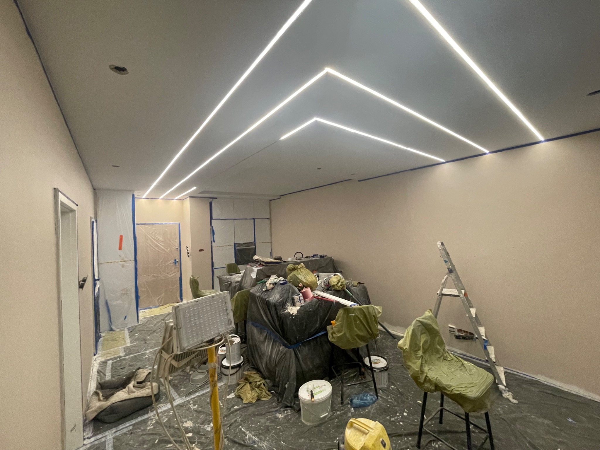 Wnętrze w trakcie malowania: ściany w beżu, zabezpieczone meble, rozstawiona drabina i lampa. Sufit z nowoczesnym oświetleniem LED w geometrycznym wzorze.