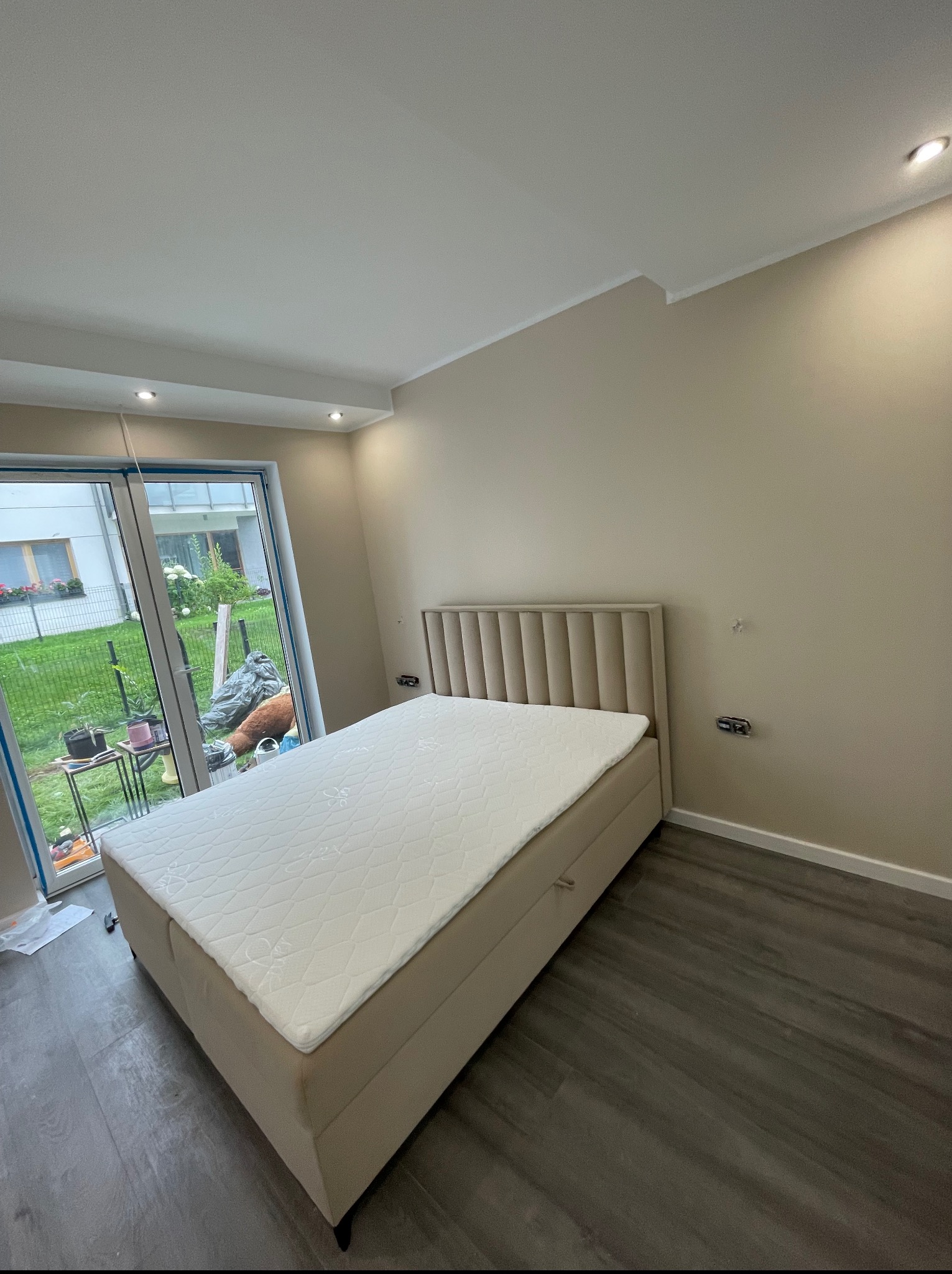 Beżowa tapeta w pokoju z łóżkiem w Mławie. Widoczne gniazdka elektryczne na ścianie, okno z widokiem na ogród i panele podłogowe imitujące drewno. Stonowana kolorystyka.