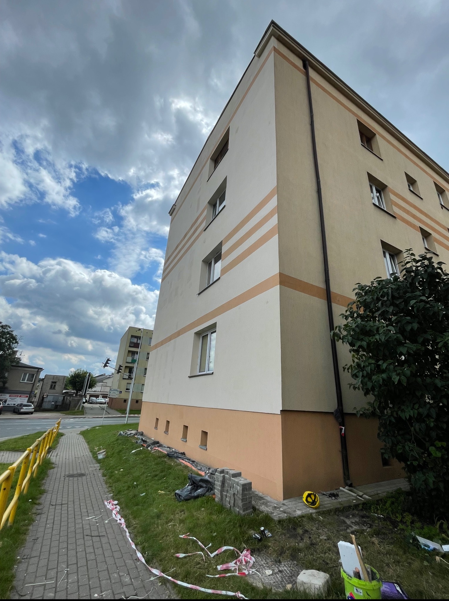 Odnowiona elewacja bloku z beżowymi pasami i oknami, widok z dołu, obok chodnik i trawnik z elementami robót wykończeniowych, niebo z chmurami.