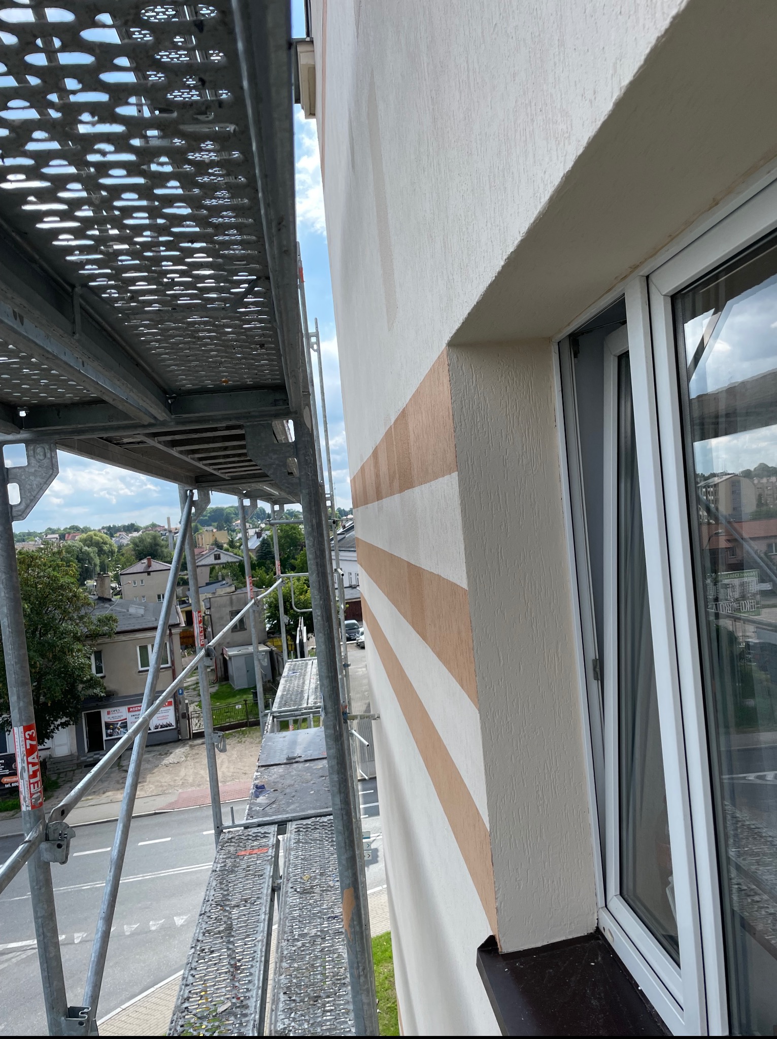 Elewacja budynku w kremowych pasach, widziana ze rusztowania z perspektywy góry. Okno z białymi ramami. W tle zabudowa miejska i zieleń.